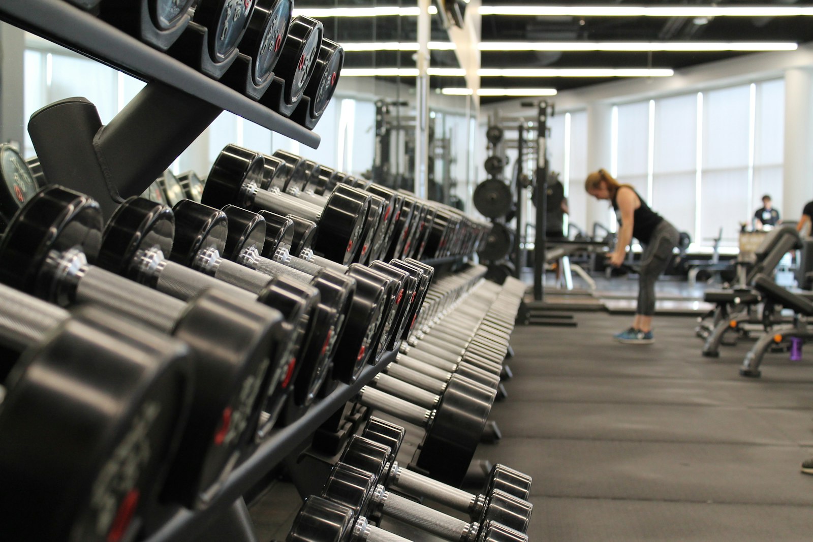 A long rack of dumbbells in a gym, with a lifter working in the background