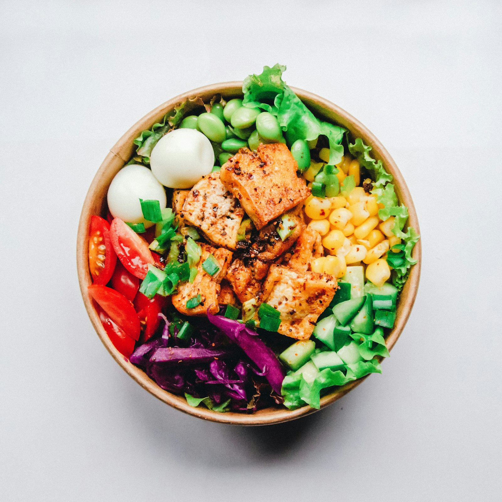 A grain bowl photographed from directly above, divided into portions of tofu, greens, corn, tomato and cucumber