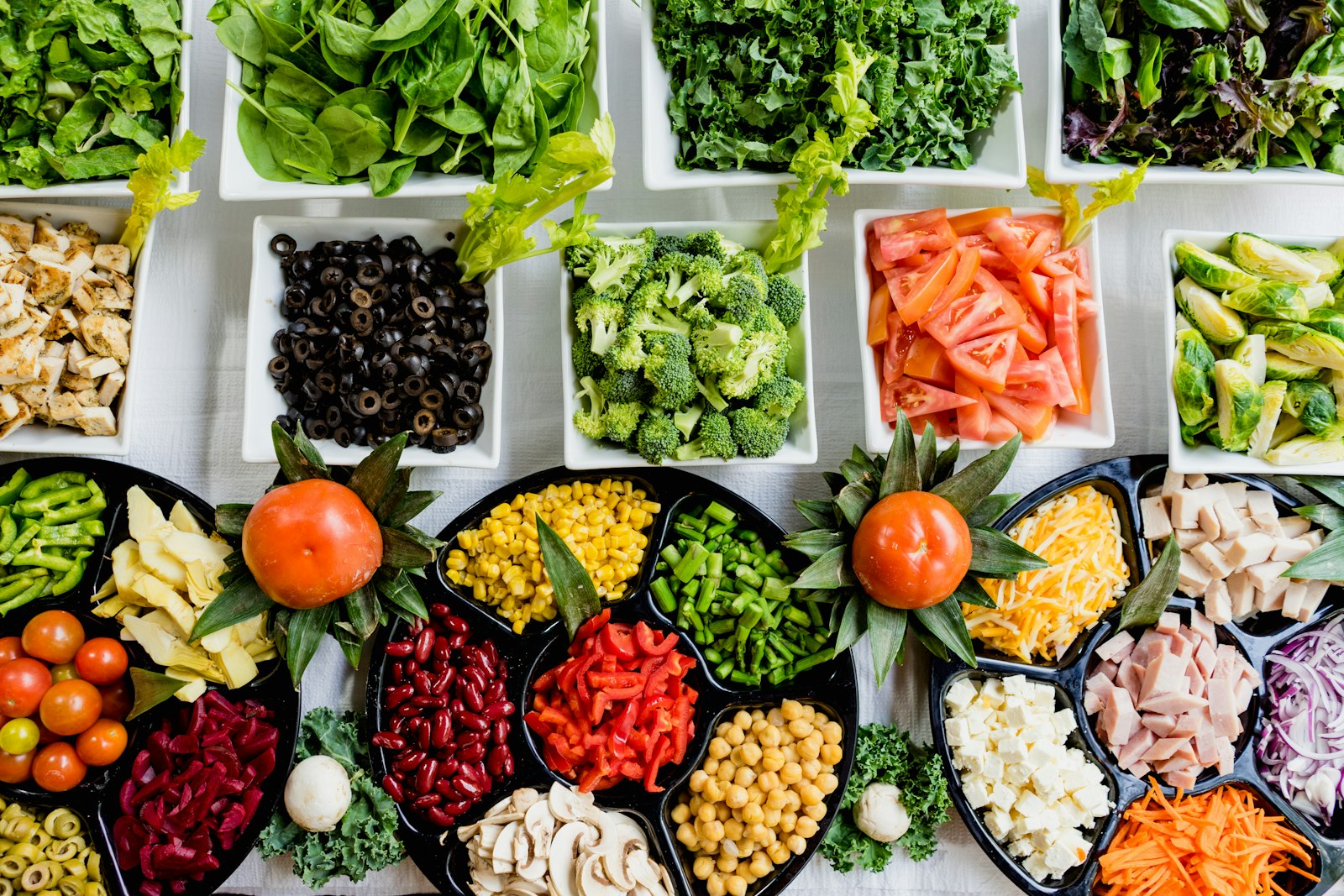 A counter of prepared salad bar ingredients in white and black trays: greens, olives, tomatoes, beans, chickpeas, cheese and shredded carrot